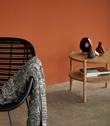Un interno elegante con il tavolino Acron di Hübsch in legno chiaro. Il tavolino rotondo è posizionato davanti a una calda parete color terracotta ed è decorato con vasi e libri. Una sedia nera con una coperta drappeggiata completa l'atmosfera accogliente e minimalista