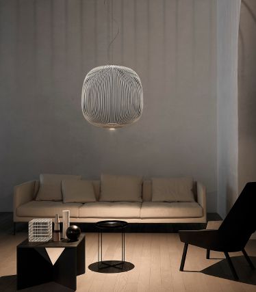 Un soggiorno minimalista con la lampada a sospensione Spokes di Foscarini come elegante punto focale. I delicati montanti metallici della lampada proiettano morbidi giochi di luce sulle pareti e sul soffitto, conferendo alla stanza un effetto leggero e scultoreo. Un divano chiaro, accenti scuri e materiali naturali creano un'atmosfera calma e moderna.