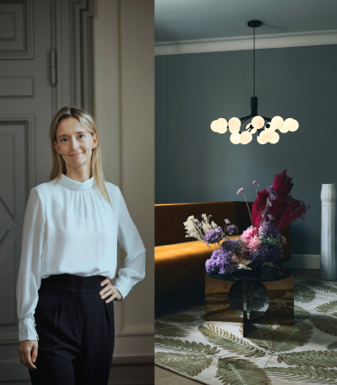 Ritratto della designer Sofie Refer accanto a una luminosa lampada a sospensione di Nuura in un interno elegante con accenti floreali.