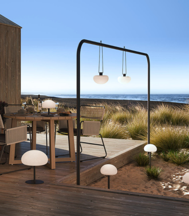 Lampade da esterno su una terrazza in legno sul mare, con luce soffusa tra l’erba delle dune.
