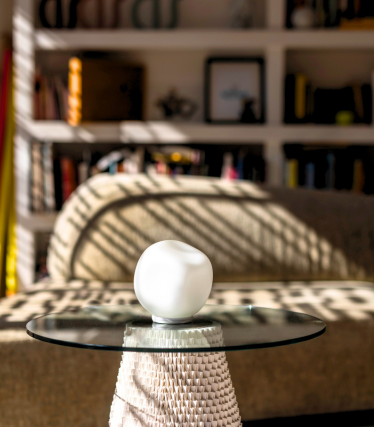 Piccola lampada da tavolo bianca su piano in vetro, con luce solare e libreria sullo sfondo.