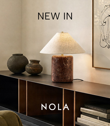 Lampada da tavolo Nola con base in marmo marrone rossiccio e paralume conico in lino su una credenza scura. Testo: «Novità – Nola».
