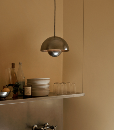 Lampada a sospensione in metallo lucido sopra una mensola con ciotole in ceramica e bicchieri, davanti a una parete color sabbia.