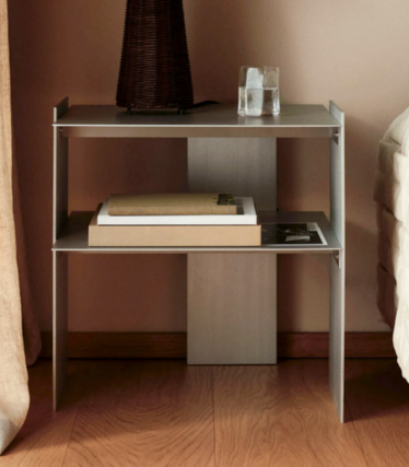 Comodino stretto in metallo con due ripiani, libri, un bicchiere e un vaso accanto al letto.