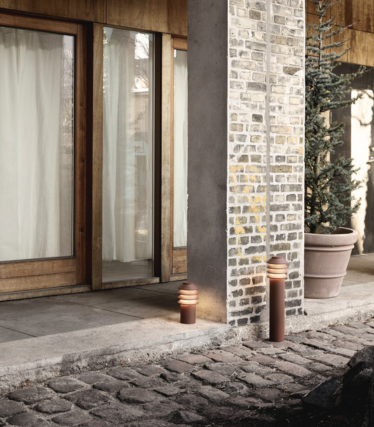 Scena esterna con due moderne luci cilindriche posizionate accanto a un vialetto in pietra e una colonna in mattoni e cemento. Dietro di esse si trovano porte in vetro con telaio in legno e tende luminose, oltre a una grande pianta sempreverde in vaso. La calda luce delle lampade crea un'atmosfera accogliente e invitante.