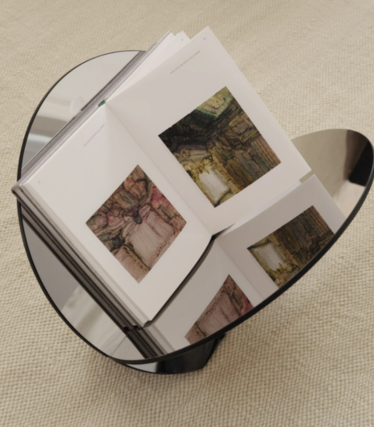Tavolino rotondo in vetro scuro con un libro d’arte aperto, su un tappeto beige.