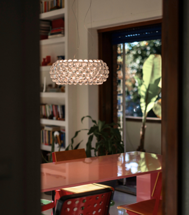 Lampada a sospensione sferica composta da sfere di vetro sopra un tavolo da pranzo rosso, vista attraverso una porta.