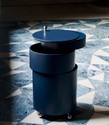 Carrello bar cilindrico blu scuro Verpan con ruote e coperchio rimovibile su pavimento in pietra decorato.