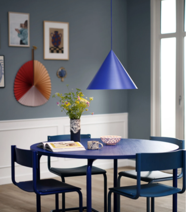 Lampada a sospensione blu a forma di cono sopra un tavolo da pranzo rotondo blu con un mazzo di fiori, in una sala da pranzo con parete blu e opere d’arte.