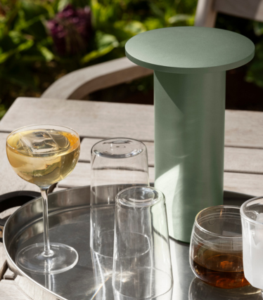 Tavolino/refrigeratore cilindrico verde salvia su un vassoio con bicchieri e bevande all’aperto.